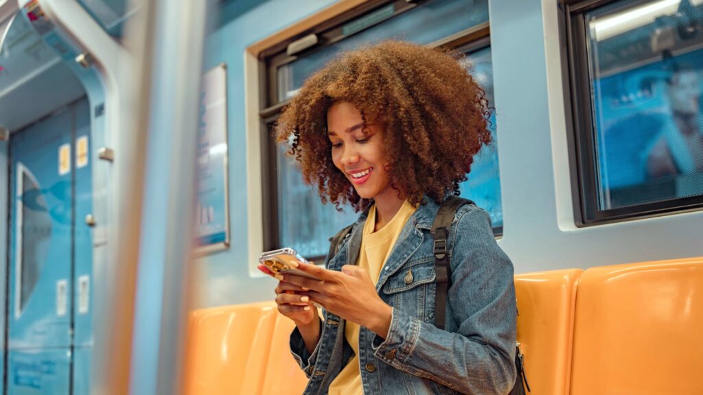 Young woman with curly hair smiling and using a smartphone on a subway train.